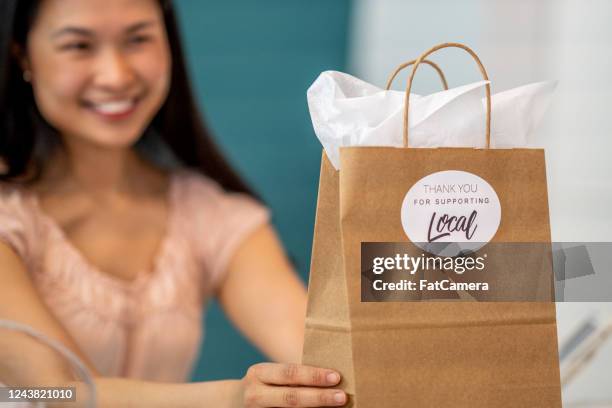 smiling female shop owner holding up a craft bag with a "thank you for supporting local" sticker - ethical consumerism stock pictures, royalty-free photos & images