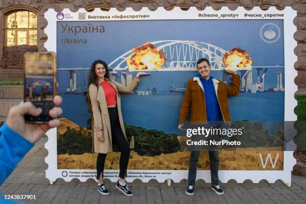 People pose for photos and take selfies in front of the large poster form of postage stamp depicting the Crimean Kerch Bridge on fire in Kyiv,...