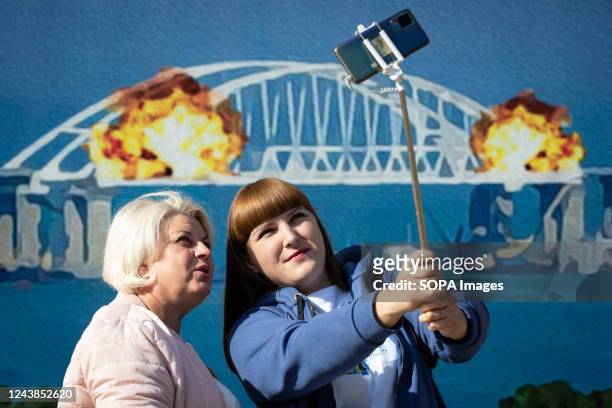 Women take a selfie with a stamp model depicting the burning Crimean bridge in the background, in central Kyiv. On October 8, an explosion occurred...