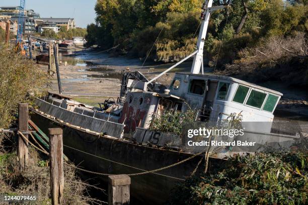 Vandalised derelict boat sits in low-tide river mud and silt at Brentford Ait, on 18th October 2022, in London, England.