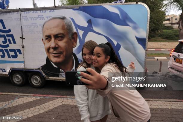 Two girls pose for a "selfie" photo before a camper wagon showing an Israeli Likud party electoral banner depicting its leader former prime minister...