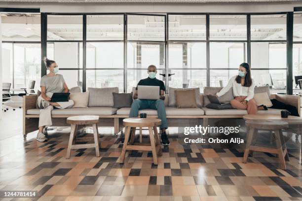 geschäftsleute, die auf einem sofa in einem modernen büro arbeiten und soziale distanz pflegen - abstand halten infektionsvermeidung stock-fotos und bilder