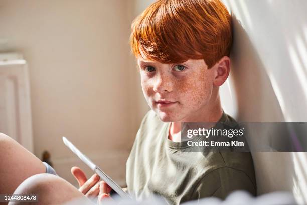 boy with red hair sitting on floor in sunny room, holding digital tablet. - red hair boy and freckles stock-fotos und bilder