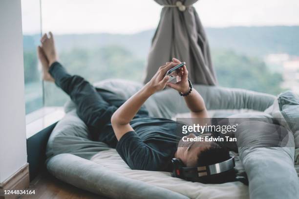 an asian chinese teenager boy listening to music with his headphone and his smart phone lying down on cushion - boy ipod stock pictures, royalty-free photos & images