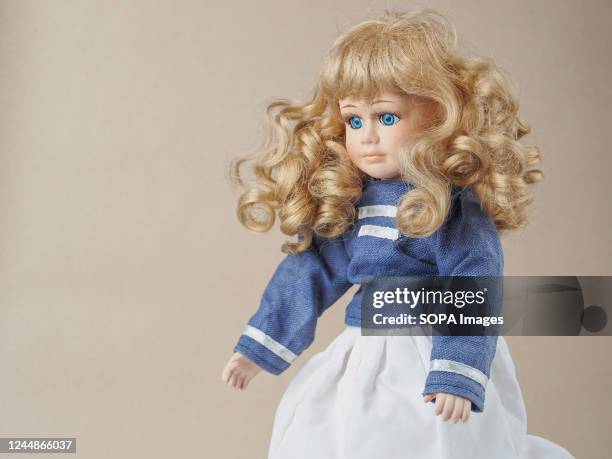 In this photo illustration, a vintage porcelain doll girl with blue eyes and blonde loose curly bare hair in a blue blouse with two white horizontal...