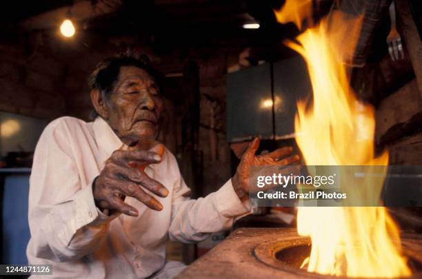 Guató indigenous people, elderly native man warms his hands at the firewood stove - acculturated indian lives in urban shantytown.