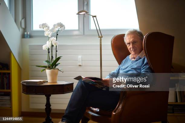 senior man sitting in armchair at home reading magazine - leather journal stock pictures, royalty-free photos & images
