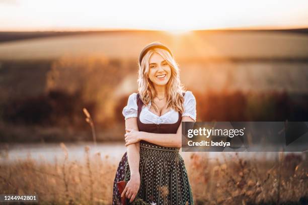 Pretty blonde girl in dirndl, standing outdoors in the field, holding bouquet of a field flowers.