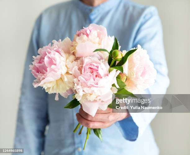 man in blue shirt holding beautiful a bouquet of peonies in tender pink and peach colors, cropped photo, bouquet close up on the light background. - close up of flower bouquet stock-fotos und bilder