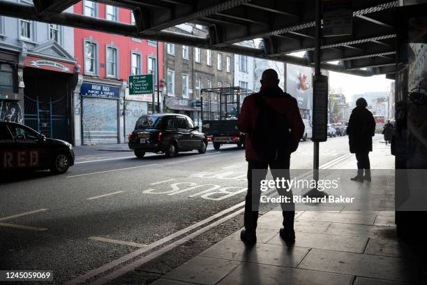 As traffic queues on the southbound carriageway, the silhouettes of commuting passengers wait for their next service north at a bus stop on the A10...