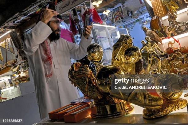 Merchant uses a phone to check his styling as he stands before golden boot trophy replicas and other memorabilia at the Souq Waqif market in Doha on...