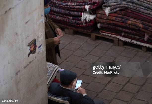 An Iranian trader uses his smartphone to check his social media pages while sitting next to handwoven rugs at a carpet market in Tehran Grand Bazaar...