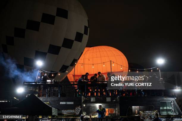 Man cooks meat on a grill as people enjoy dinner on a terrace in front of hot air balloons displayed as part of the Expo Transporte Internacional...