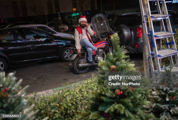 An Iranian trader wearing a Christmas fun hat while speaking on his smartphone as he sits on a motorcycle on a street-side in downtown Tehran,...