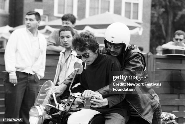 American singer Bob Dylan, in sunglasses and with a cigarette in his mouth, and folk musician Bob Siggins, in white helmet, ride a motorcycle...
