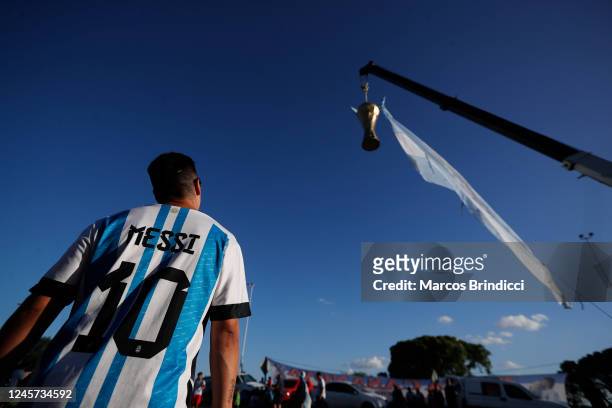 Fan wears a jersey of Lionel Messi watches a large replica of the FIFA World Cup hanging from a crane before the arrival of the Argentina men's...