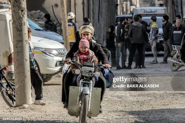 Syrian man and a child ride on a motorcycle bearing the image of Iraq's late president Saddam Hussein on the headlight, as rescue teams inspect the...