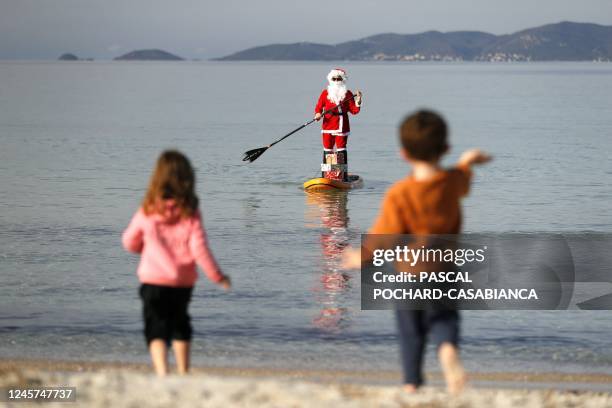 Children wave at a woman dressed as Santa Claus steering a paddleboard close to the Mare e Sol beach in Pietrosella on December 20, 2022 on the...