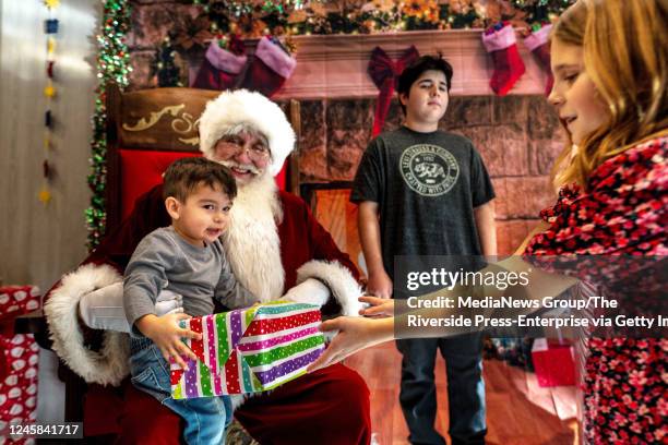 Temecula, CA Aiden Lamas receives a Chrismas present from volunteer, Lucy Deel right, as he sits with Santa Clause during the 28th annual free...