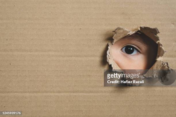 a child eye looking through a hole on a cardboard box. concept of spy and stalker - neugierde stock-fotos und bilder