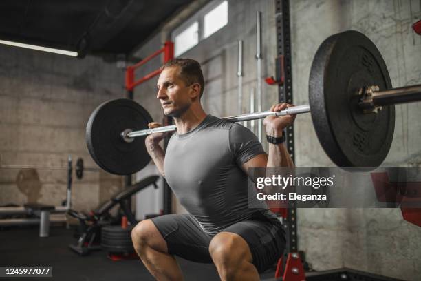 handsome muscular man doing squat exercise with barbell at the gym - crouch stock pictures, royalty-free photos & images