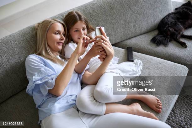 happy mother and daughter sharing smartphone on couch in living room at home - woman smartphone family ストックフォトと画像