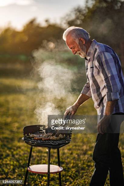 happy senior man grilling meat on barbecue in nature. - barbecue man stock pictures, royalty-free photos & images