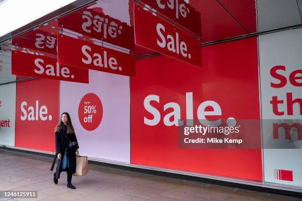 People out shopping on Oxford Street walk past large scale January sale signs in red and white for major high street clothing retail shops including...