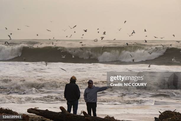 Large waves crash behind two men on Rio Del Mar Beach, covered in storm debris, in Aptos, California on January 12, 2023. - A "relentless parade of...