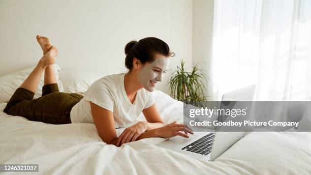 smiling woman in a face mask lying on her bed using a laptop - pleased face laptop stock pictures, royalty-free photos & images