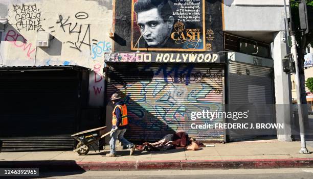 Construction worker pushes his wheelbarrow past a homeless man sleeping on a Hollywood sidewalk on January 20, 2023 in Hollywood, California. - Los...