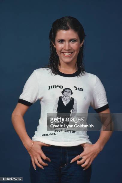 Portrait of American actress Carrie Fisher, dressed in jeans and a t-shirt, as she poses against a blue background, 1978. Her t-shirt advertises...