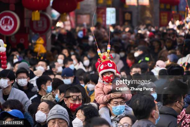 Among the crowded people in a temple fair on Tianjin ancient cultural street, a little girl wearing mask and traditional hood is sitting on her...