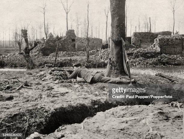 British sniper on duty lying alongside a riverbank amidst the ruins of a village near Verdun on the Western Front during World War One, circa 1916.