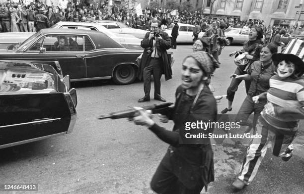 Female demonstrators, including one with a toy gun and another in costume, run towards the camera while participating in Vietnam War protests, with...