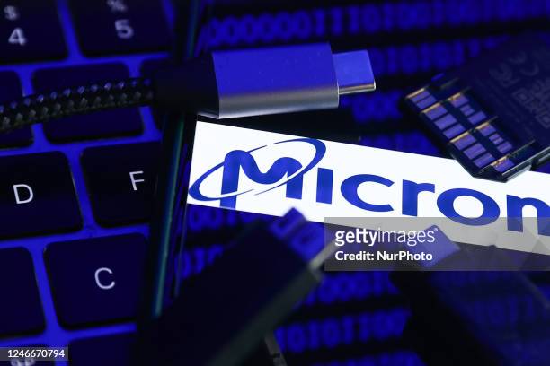 Micron logo displayed on a phone screen with a binary code reflected on it, a laptop keyboard, a memory card, an adaper and cables are seen in this...
