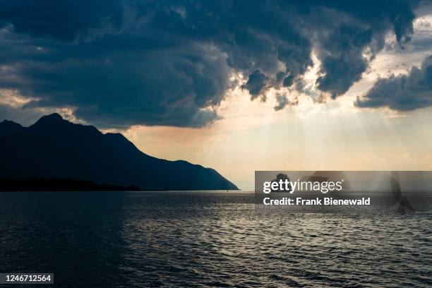 Dark storm clouds building up over Lake Geneva, a single tree on a small island and alpine scenery in the distance.