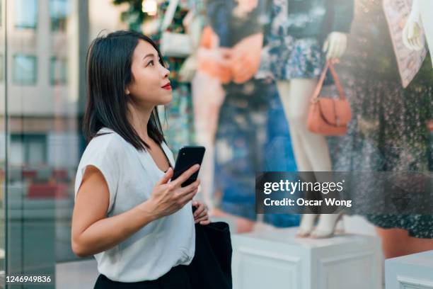 young asian woman window shopping with smartphone on the high street - woman shopping china stock pictures, royalty-free photos & images