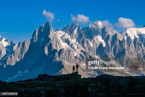 The peaks of the Mont Blanc massif are reflecting on the surface of Lacs des Chéserys, two hikers are standing on a ridge in the distance.