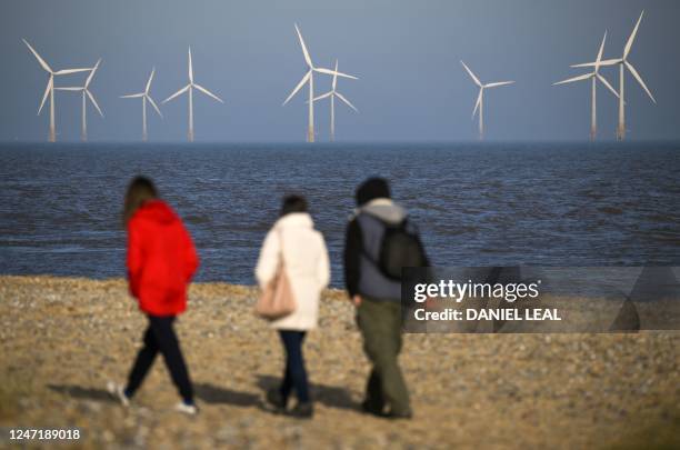 Pedestrians walk along the shore as wind turbines are pictured at RWE's Scroby Sands Wind Farm, off the coast of Great Yarmouth, eastern England, on...
