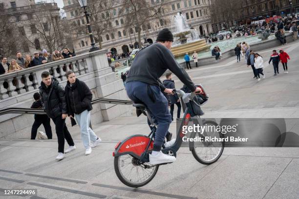 Santander rental bike user bumps down steps in front of visitors and tourists to Trafalgar Square, on 17th February 2023, in London, England.