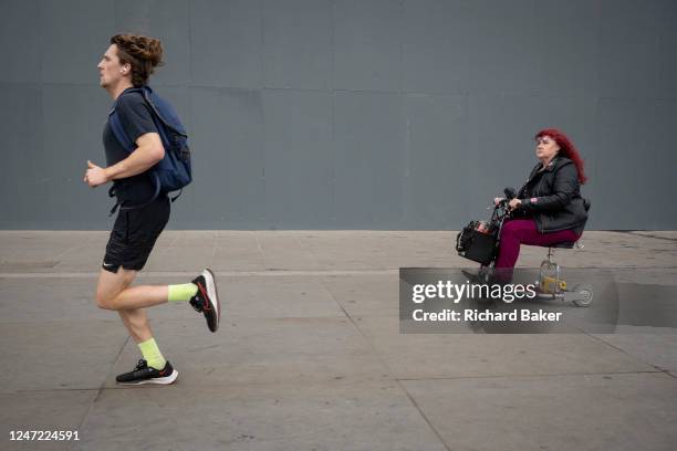 Runner speeds past a woman seated in a small mobility scooter, in front of the National Gallery which is currently undergoing extensive refurbishment...