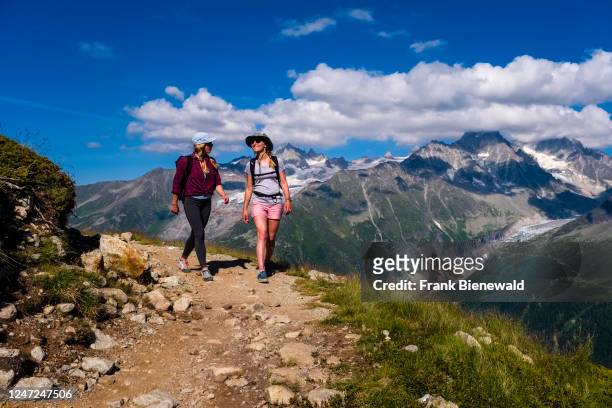 Two young female hikers walking on a path near La Flégère, the peaks of the eastern part of the Mont Blanc massif in the distance .