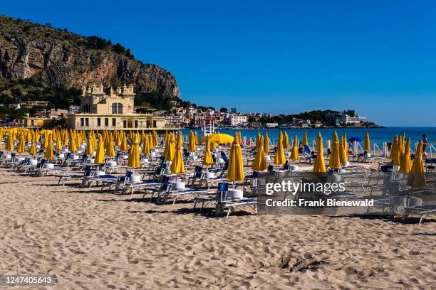 Empty sun loungers and yellow parasols set on the beach of the small town of Mondello, the rock face of the mountain Mt. Gallo behind the houses.
