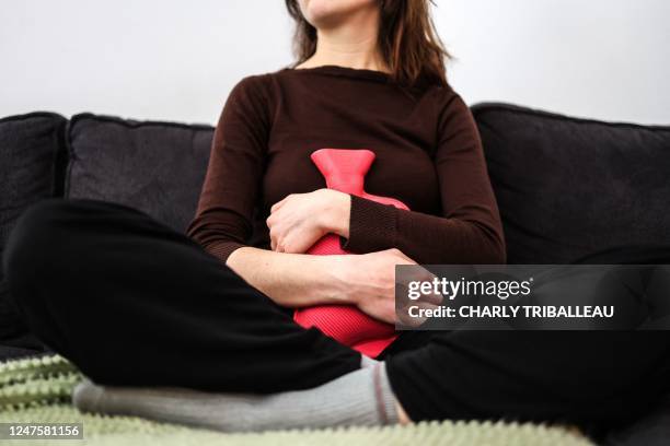 This illustration photograph taken on March 1, 2023 shows a woman with painful periods sitting on a sofa while holding a hot water bottle in...