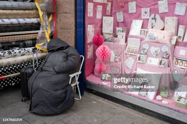 Homeless person sits in a chair on the high street in Cricklewood, outside a retailer selling a pink-themed celebration cards, on 6th March 2023, in...