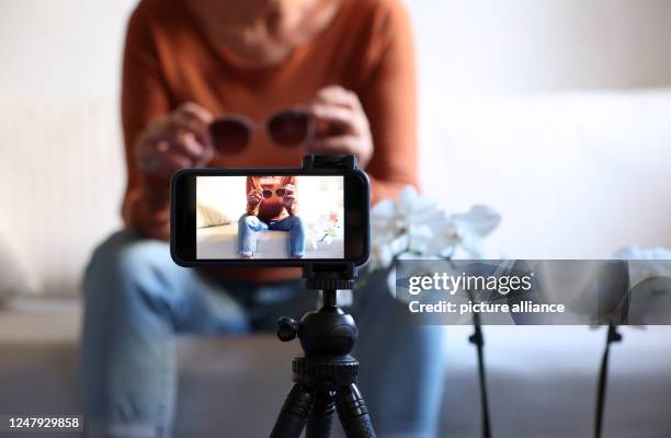 March 2023, Bavaria, Kaufbeuren: A woman sits on a sofa with sunglasses in her hands while filming herself with her iPhone . Product tips on...