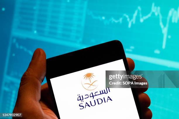 In this photo illustration, the Saudi Arabian Airlines logo is seen displayed on a smartphone with an economic stock exchange index graph in the...