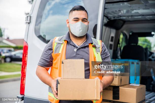 junge männliche hispanische lieferung mann nimmt paket nach hause tragen eine gesichtsmaske - essenzielle berufe und dienstleistungen stock-fotos und bilder