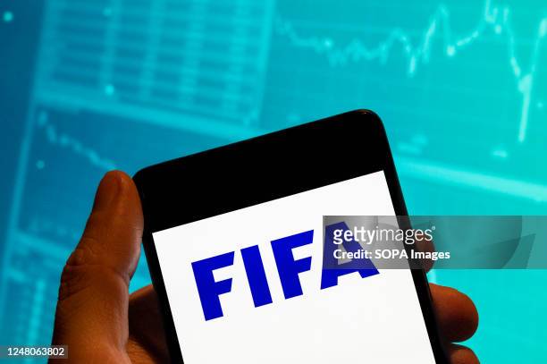 In this photo illustration, the Fédération Internationale de Football Association logo is seen displayed on a smartphone with an economic stock...